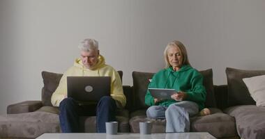 An old European man uses a laptop while sitting on a sofa in a modern home setting next to an older woman using a tablet. Modern pastime of an elderly couple video