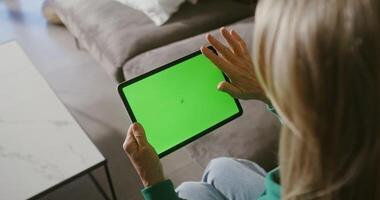An elderly woman uses a tablet with a green screen, holding it in front of her and sliding her fingers across the screen. Copy space, chroma key, woman's face is not visible, filming from behind video