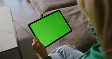 An elderly woman is chatting on a tablet with a green screen, holding it in front of her. Copy space, chroma key, woman's face is not visible, filming from behind video
