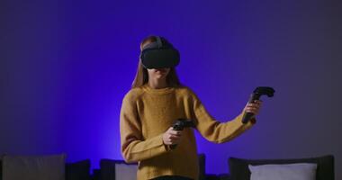 Virtual reality, online game. Young beautiful girl in virtual reality glasses with joysticks in hands slowly moves in a stylish home interior with a wall illuminated in blue video