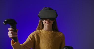 Virtual reality, online game. Young beautiful girl in virtual reality glasses with joysticks in hands slowly moves while standing in front of a plain blue wall video