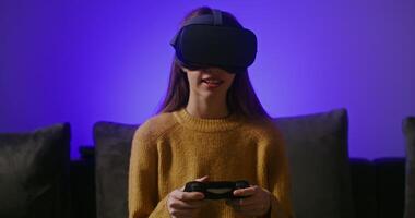 A cute teenage girl dressed in casual clothes in large virtual reality glasses, plays an online game, sitting on the couch with a joystick in her hands, against the background of a blue wall video