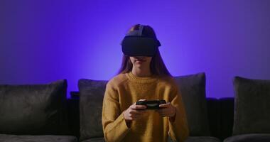 A girl of European appearance, dressed in casual clothes in large virtual reality glasses, smiling while playing a computer game, sitting on a sofa with a joystick in her hands video