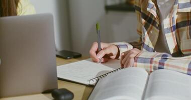 A young guy of European appearance makes notes in a notebook while doing homework paired with a girl sitting at an open laptop, close-up of a writing hand, people's faces are not visible video