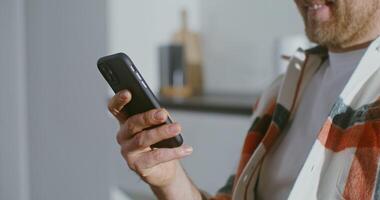 Young fair-haired bearded man of European appearance smiles while looking at the screen of a mobile phone, sitting at the table in the kitchen. Close-up filming from the side video