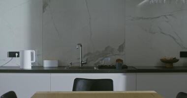 Young wide-awake male european appearance includes a kettle, and sit down at the table in a modern stylish kitchen with a phone in his hands waiting for a kettle boiling video