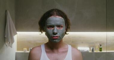 Young curly-haired man of European appearance with a clay mask on his face smiling while looking straight into the camera, portrait shooting in a modern bathroom video