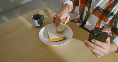 A man dressed in casual clothes is having breakfast while sitting at the table in the kitchen and uses a mobile phone, a close-up of the set table and the man's hands, man's face is not visible video