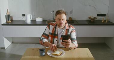 Young bearded man of European appearance does not take his eyes off the screen of a mobile phone while having breakfast, in a modern stylish kitchen video