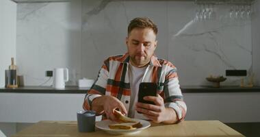 A young bearded man of European appearance drinks coffee and eats a sandwich without taking his eyes off mobile phone screen in his hand, laughing at what he saw, sitting at table in the kitchen video
