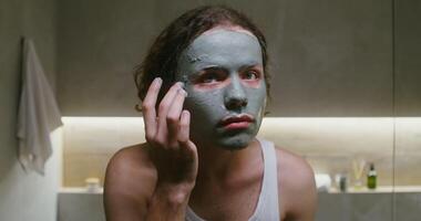 Young curly-haired man of European appearance with a clay mask on his face looks at the camera as if in mirror, touching up the clay on his face with his fingers, front camera, portrait videography video