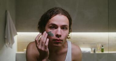 Young curly-haired man of European appearance smears a clay mask on his face, looking directly into the camera as in a mirror, portrait in a modern bathroom video