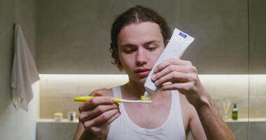 Young curly-haired man of European appearance smears toothpaste on a toothbrush and begins to brush his teeth, looking directly into the camera as in a mirror, standing in a modern bathroom video