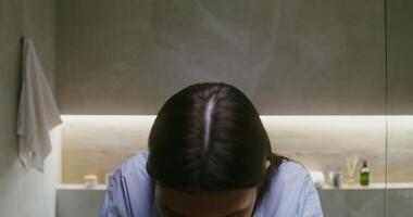 A young beautiful woman of European appearance, dressed in a loose shirt, washes her face with water and then dries it, patting it with a towel, looking at the camera as in a mirror video