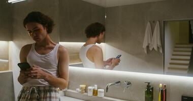 A young curly-haired man of European appearance, waiting for the action of patches applied to their eyelids, uses a mobile phone, leaning on the sink, standing in a modern stylish bathroom video