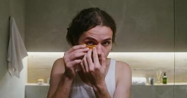 Young curly-haired man of European appearance puts patches under his eyes, looking at the camera as in a mirror, frontal view, portrait in a stylish bathroom video