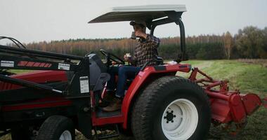 Young black haired male farmer eating a sandwich and drinking tea from a metal mug, while sitting in the open cabin of a modern agricultural tractor parked in the middle of a field for lunch video