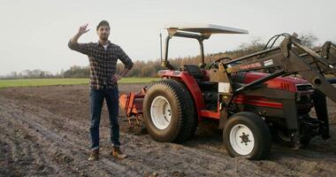 A young male farmer of European appearance, straightens his cap while smiling and looks directly into the camera, standing next to a small universal agricultural tractor in a field on an autumn day video