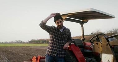 A young male farmer of European appearance straightens his cap while smiling and looking directly at the camera, leaning on a small agricultural machine standing in a field video