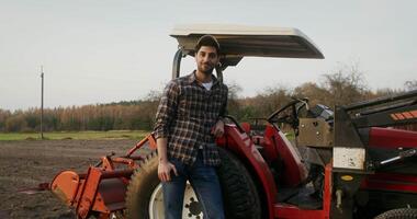 A young male tractor driver of European appearance stands leaning on an agricultural machine and smiles while looking directly into the camera, in a field on an autumn day video