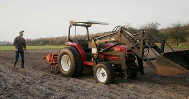 A young man of European appearance in a cap, walks through a plowed field to a tractor and sits on the seat of an open cabin behind the wheel video