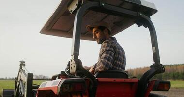 A young European farmer operates a farm tractor, while sitting in an open cab with canopy and turning back to control the evenness of the plowing strip video