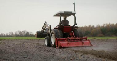 A young male farmer in a cap drives a tractor across the field, plowing it with a plow, and turning back to check his work. filmed from behind video
