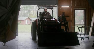 A young man of European appearance with a beard, starts a small agricultural tractor with a bucket and drives it out of the garage, backing it up, going to work on the farm on an autumn day video