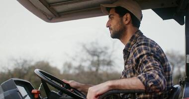Young dark-haired male farmer with a beard, with a cap on his head, rides a tractor with an open cab, looking around, inspecting his property, close-up video