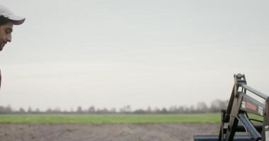 A young black-haired male farmer with a beard gets behind the wheel of a small farm tractor and starts plowing through the field. filming from behind, close-up video