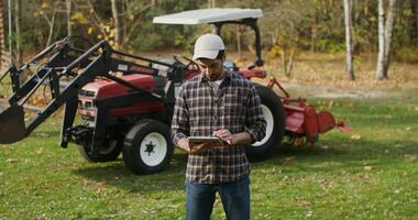 Young man of european appearance in baseball cap smiling broadly looking straight at the camera after using the tablet, while standing near a modern tractor on an autumn day video