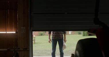 A young handsome man with a beard enters the garage after waiting for the gate to open and, putting a cap on his head, begins to inspect an agricultural vehicle parked in the garage video