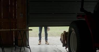 A young black-haired man with a beard opens the garage door and walks inside, approaching a tractor parked there, intending to work on a farm on a sunny autumn day video
