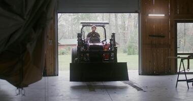 A young man of European appearance with a beard drives a small agricultural vehicle with a bucket in front into a wooden garage, about to park after work on a farm on an autumn day video