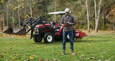 A young man of European appearance in a baseball cap, uses a tablet, standing near a modern agricultural machine on a sunny autumn day video
