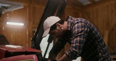 Mechanic - a young dark-haired man with a beard fixes a small farm tractor, by repairing it with tools in a wooden barn video