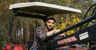 A young man of Caucasian ethnicity in a baseball cap, sits with his hands on the steering wheel of an agricultural car and smiling while looking at the camera on a sunny day video