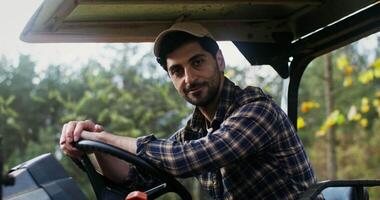 Young black-haired man of European appearance with a beard smiling, while looking at the camera while sitting at the wheel of an agricultural car with a canopy video