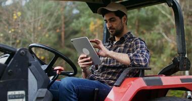 A young dark-haired man of European appearance in a baseball cap uses a tablet, while sitting in a standing agricultural machine on a farm on a sunny autumn day video