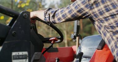 Young bearded dark-haired man in a baseball cap climbs onto a tractor seat, getting ready for a trip, close-up video