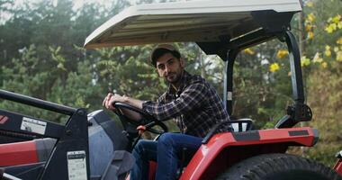 Young black-haired man with a beard with a baseball cap on his head smiling, while looking at the camera while sitting at the wheel of an agricultural car video