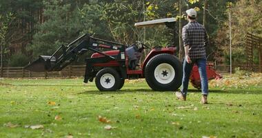Young black haired man wearing baseball cap walks up, sits down, turns on and starts driving a modern small farm tractor with a bucket in the front and a plow in the back video