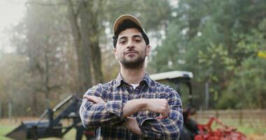 Young dark haired male farmer with stubble on his face smiles while looking straight into the camera and crossing his arms over his chest, as he stands near a modern agricultural machine on a ranch video