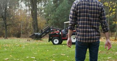 A young black-haired man with a beard, dressed in casual clothes with a baseball cap on his head, walks up to the tractor and gets into it, getting ready to work in the field video