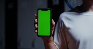 A young man with a racket in his hand holds a phone with a green screen, standing in the locker room of the sports hall. Close-up phone, man's face is not visible, copy space video