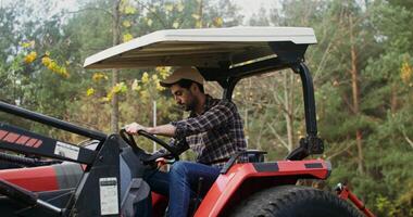 A young black-haired man with a beard, dressed in casual clothes with a baseball cap on his head, parked a tractor at the edge of the forest and got out of it, tired after working in the field video