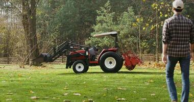 A young dark-haired man in a baseball cap walks up to a modern tractor parked at the edge of a farm and gets into it, getting ready to work in a field on a sunny autumn day video
