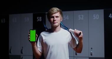 A young man of European appearance, with a tennis racket in his hands, smiling, looking straight into the camera and holding a phone with a green screen in his hands, standing in a stylish locker room video