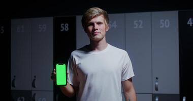 A young man, dressed in a sports uniform, smiling while looking at the camera and holding a phone with a green screen in his hands while standing in a stylish locker room. Copy space, chroma key video