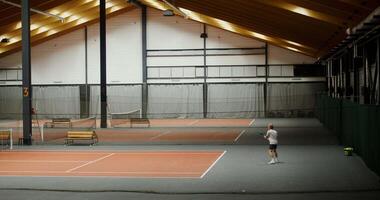 A young man of European appearance plays on one of the tennis courts in the indoor gym, actively moving and hitting the ball with a racket. Filming from afar video